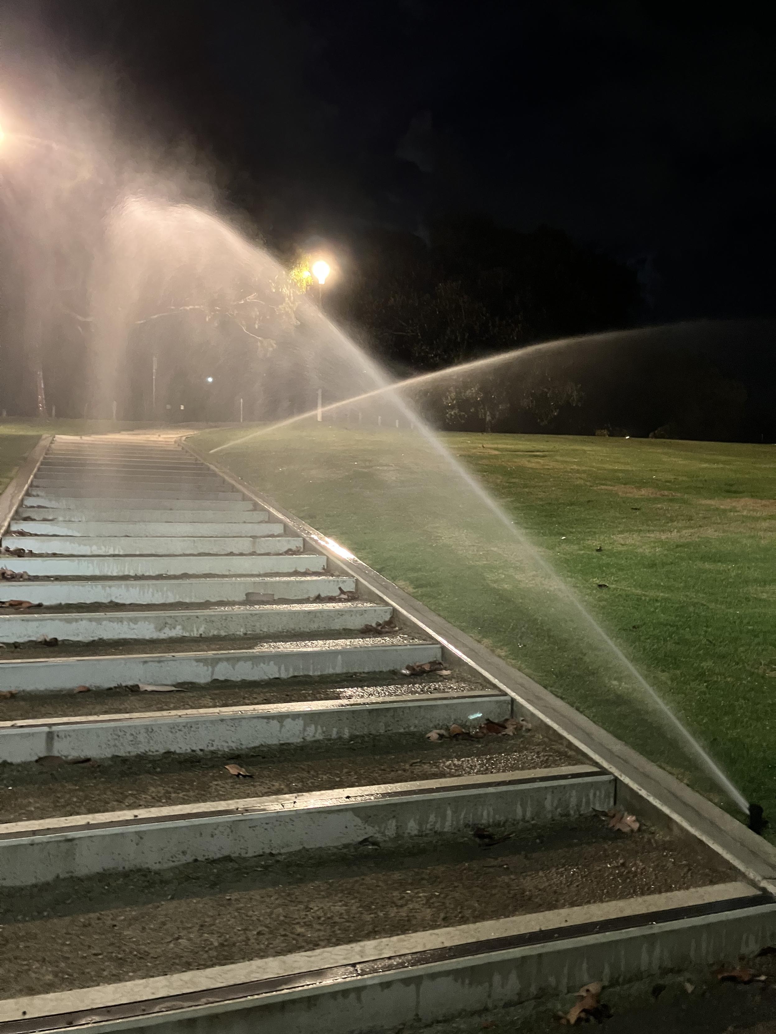 Some water sprinklers spraying a staircase next to a grassy hill. 

It’s pre dawn.