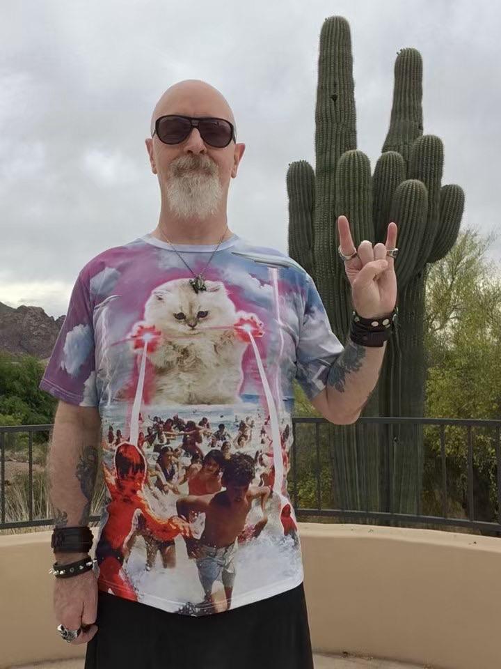 A picture of Rob Halford. He has a bald head and large white beard. He is wearing sunglasses.

He his holding his left hand up in the 'metal horns' salute, with a cactus in the background.

He is wearing a shirt featuring a giant cat shooting lasers out of its paws while people run away from it.