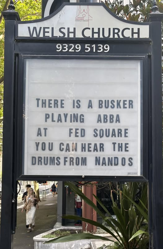 Church sign with a black frame. It's for the Welsh Church in Melbourne.
Text on the sign reads "There is a busker playing Abba at Fed Square. You can hear the drums from Nandos"