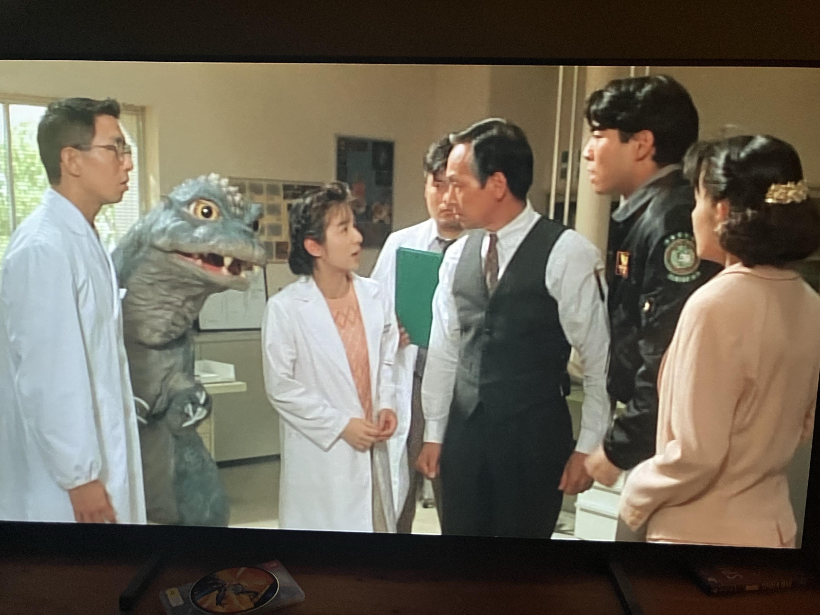 A group of 6 humans stand around to discuss something important and perhaps scientific.

Also, Godzilla Jr is trying to follow along.
